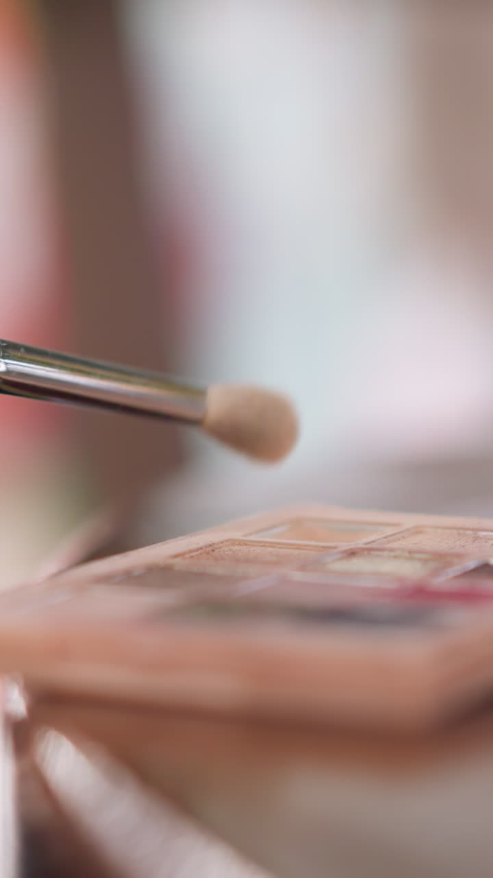 Closeup womans hand applying eyeshadow palette, soft natural light, neutral shimmer shades picked with fluffy brush, slow motion brush strokes, manicured nails, selective focus with bokeh, tutorial