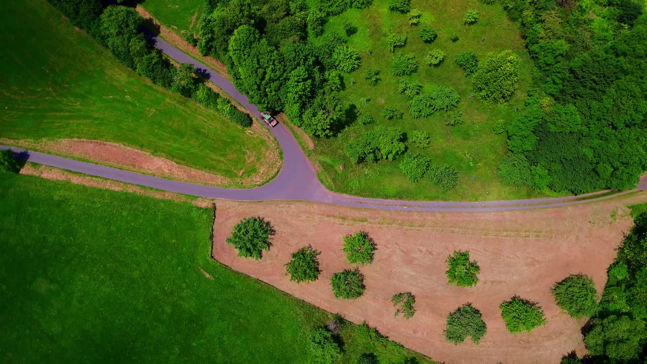 Aerial View of Small Tractor Driving on Winding Rural Road Between Green Fields and Forested Countryside in Summer