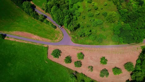 aerial view of small tractor driving on winding rural road between green fields and forested countryside in summer