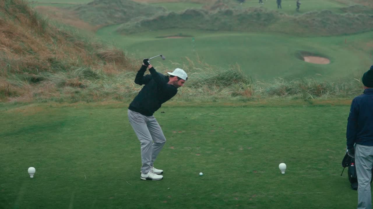 Male golfer with backwards white hat hits ball off tee from white tees on par three on a rainy foggy day on Ireland links golf course as other golfers watch on in slow motion