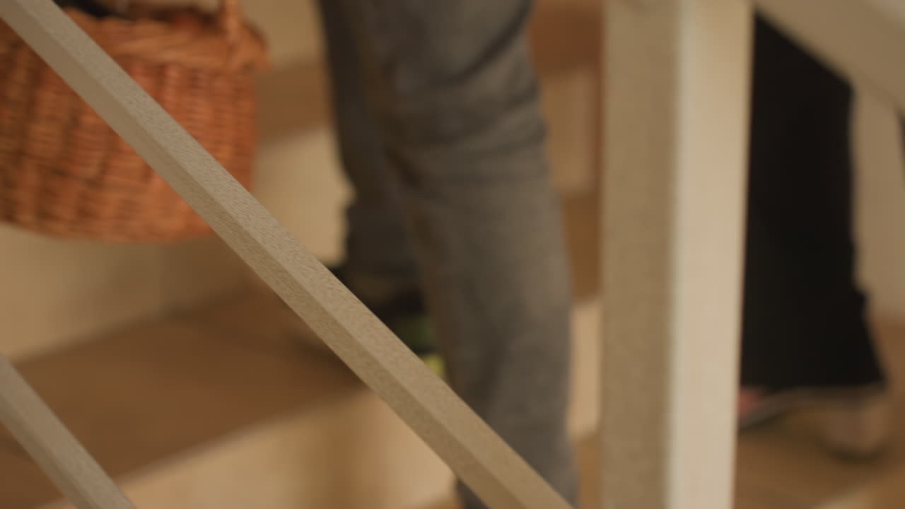 Coppia che trasporta un cesto da picnic al piano di sopra, cesto di vimini contenente una bottiglia e un regalo incartato, jeans casual e scarpe da ginnastica che salgono le scale piastrellate in un ambiente caldo e illuminato, primo piano sulle gambe e sulla mano che afferra