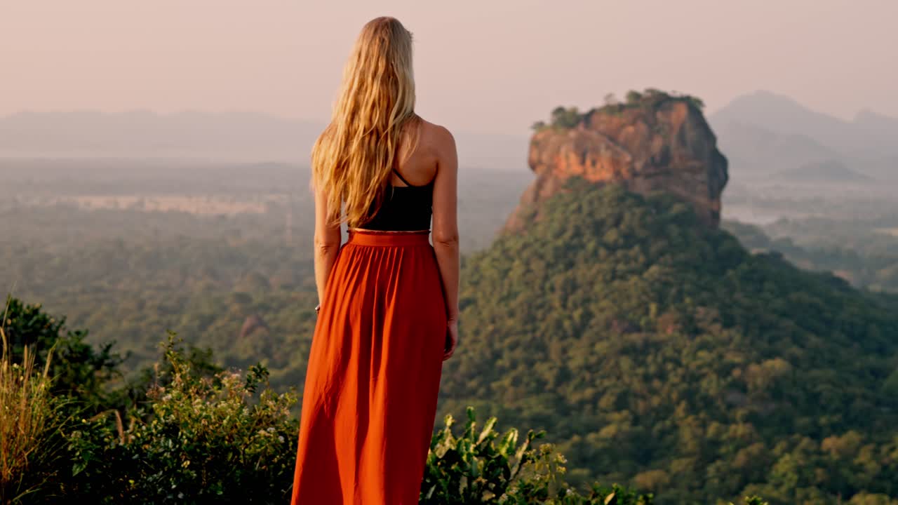A lone woman stands amid lush greenery, gazing toward the ancient Sigiriya Rock fortress as the sun rises over Sri Lanka.