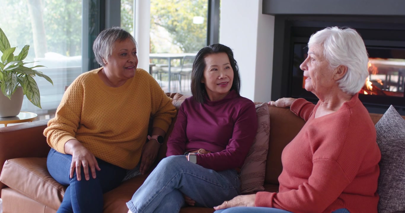 Diverse female friends sitting on sofa at home, coral sweater talking, gesturing, others nodding