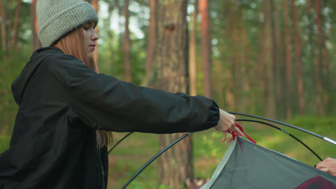 young woman in gray beanie and black jacket carefully adjust flexible tent pole during forest camping setup surrounded by trees under natural morning light focused on connecting parts securely
