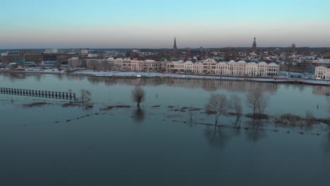 aerial cityscape approach of dutch hanseatic medieval tower town zutphen in the netherlands with snow on the boulevard at sunset reflecting with vegetation rising above overflown river ijssel