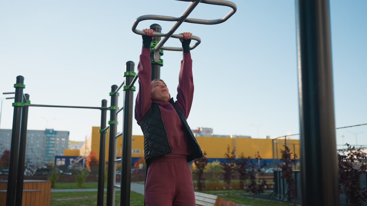 Instructor jumps on pull up bars with dynamic strength and energy, showcasing fitness training techniques in outdoor urban park under clear sky, hoodie and vest outfit framing focused action