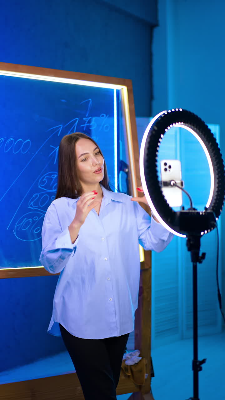 Positive brunette lady stands between glass board and phone on tripod. Blogger footages video content and farewells with audience. Vertical video.
