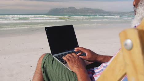 Hombre-Mayor-De-Raza-Mixta-Sentado-En-Una-Tumbona-Y-Usando-Una-Computadora-Portátil-En-La-Playa