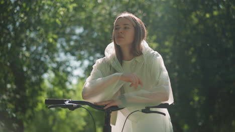 young woman in translucent raincoat removes her hood, leans thoughtfully on bicycle handlebars with hand resting on chin, sunlight softly filters through tree leaves