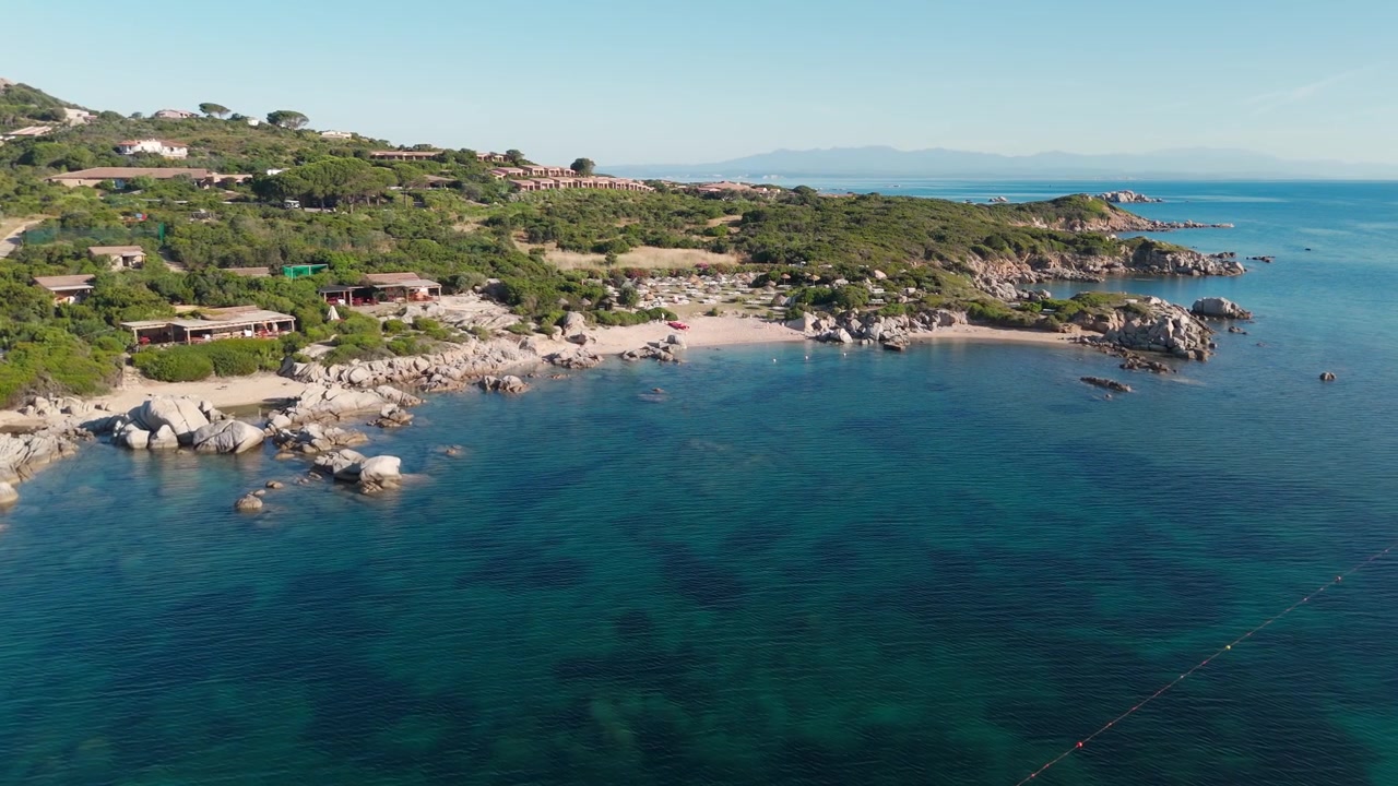 Vista aerea di costa rocciosa, mare blu cristallino e ville lungo una baia sarda.
