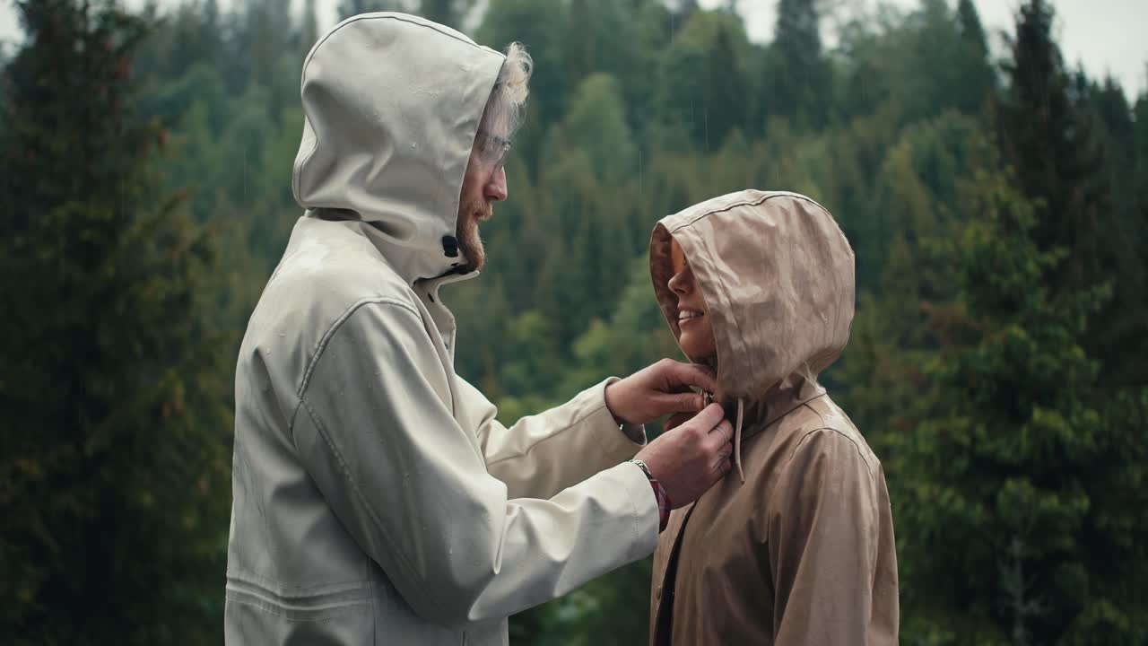 The guy fastens the girl's jacket and kisses her during the rain in a mountain coniferous forest. Happy couple against green forest background