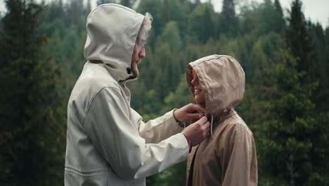 the guy fastens the girl's jacket and kisses her during the rain in a mountain coniferous forest. happy couple against green forest background