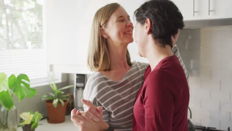 Mujer-Caucásica-Besando-A-Su-Esposa-En-La-Frente-Mientras-Bailan-Juntos-En-La-Cocina-De-Casa