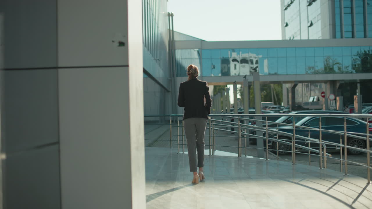 Back view of woman in professional outfit walking past modern residential building with glass facade and parked cars surrounding area in daylight, with shadows cast on clean polished walkway surface