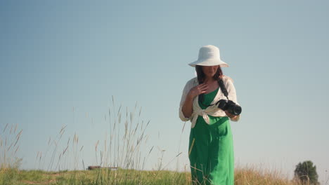 woman in green dress and white hat stands on grassy hill holding camera, enjoying peaceful nature exploration under blue sky as she prepares to capture photographs