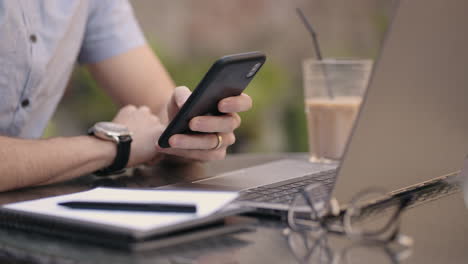 Medium-shot-of-young-man-wearing-streetwear-sitting-in-restaurant-or-cafe-in-front-of-laptop-and-having-mobile-phone.-Breakfast-With-Smartphone