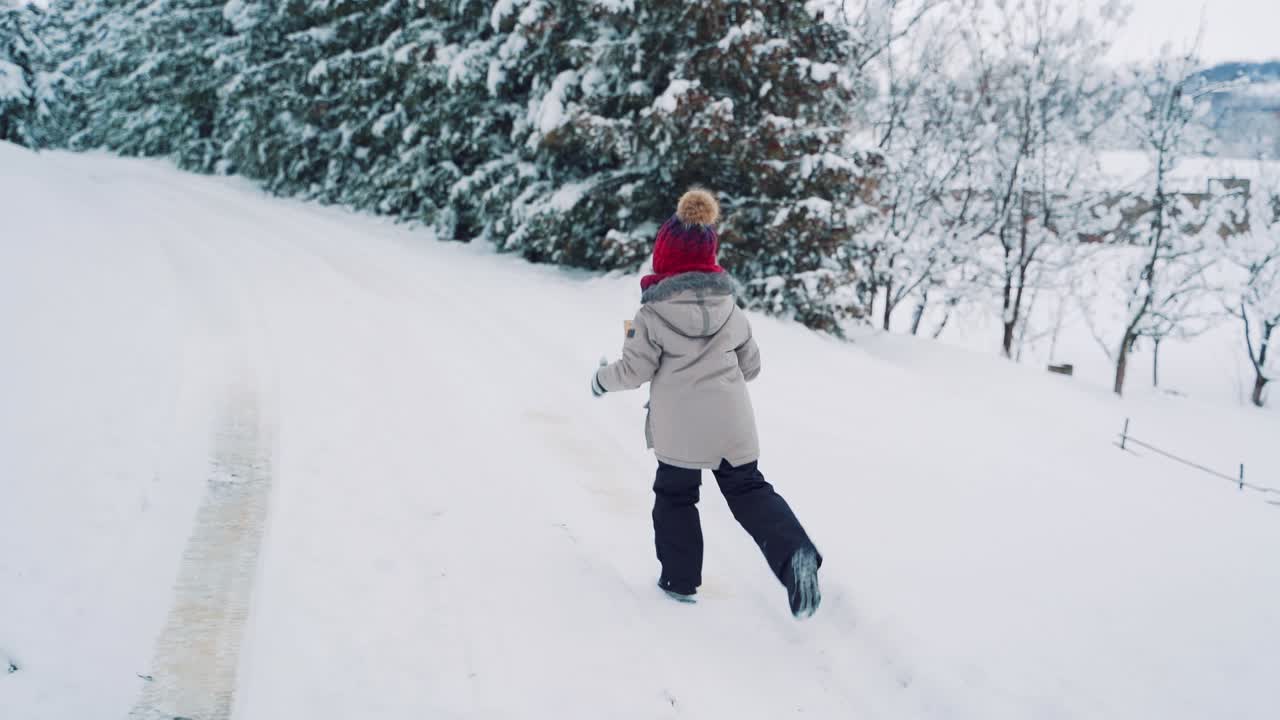 A little boy with his back in red hat is running with an envelope in his hand along a snowy path in the winter near a row of fir-trees. Slow motion