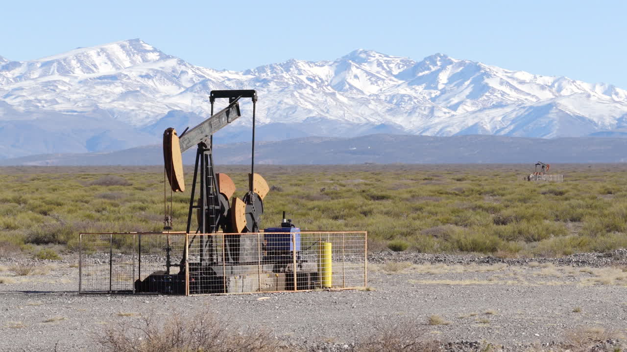 Side drone shot of oil pumpjack in Vaca Muerta with snowy Andes mountains in the background.