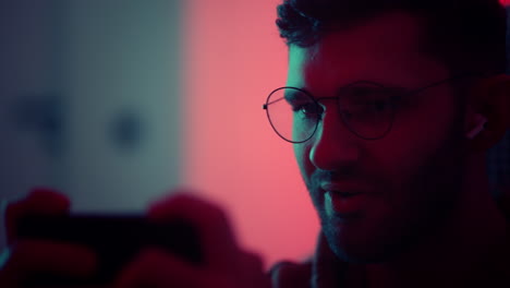 joven con gafas jugando a videojuegos con su teléfono inteligente retrato en primer plano en casa escuchando sonido con auriculares en el oído jugando en línea y transmitiendo deportes cibernéticos y ocio en su tiempo libre