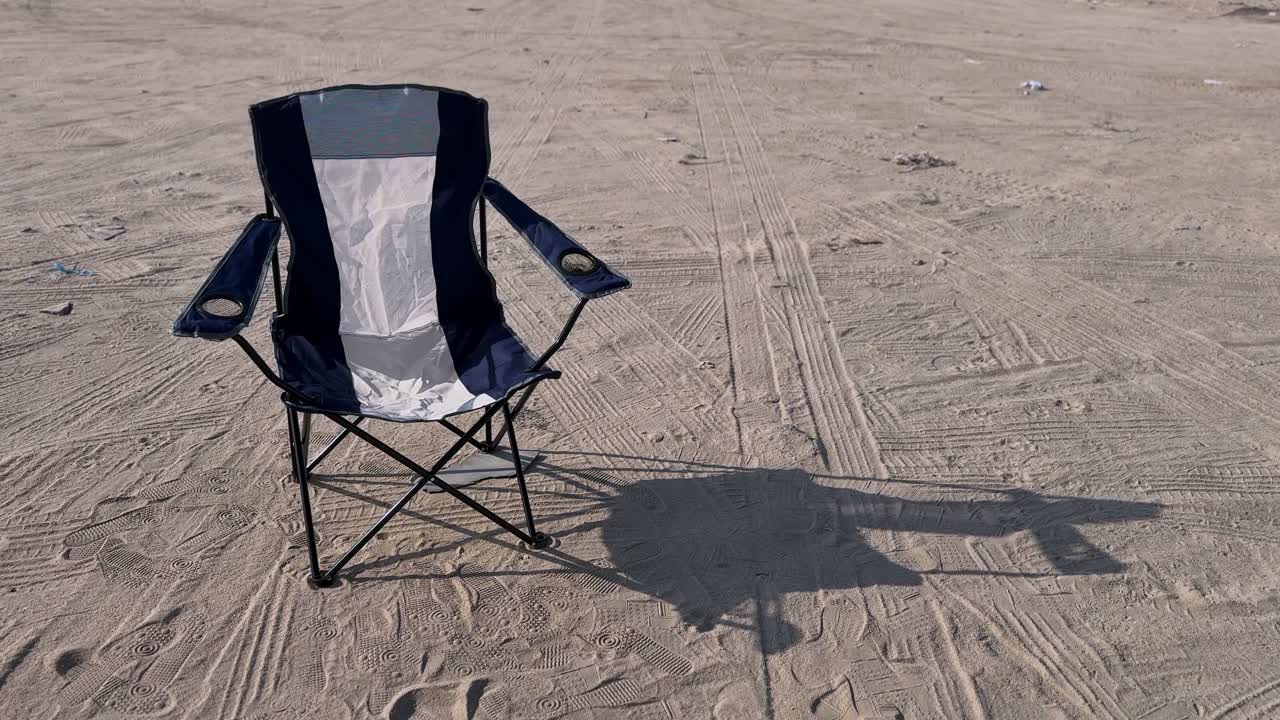 An empty blue and white folding camping chair sits alone on sandy ground covered in tire tracks and footprints. A solitary outdoor scene depicting a beach trip or desert camping adventure