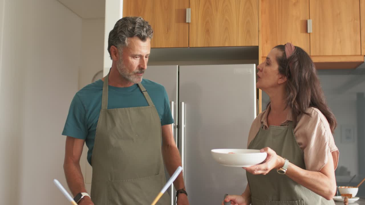 Mature couple in olive-green aprons plating at kitchen counter after cooking in skillet, serving