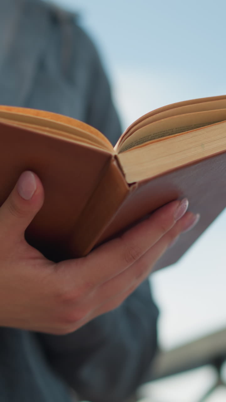 primer plano de una mujer sosteniendo un libro abierto con su mano izquierda apoyándolo desde abajo, de pie cerca de una valla de hierro, las páginas se levantan ligeramente como si de una suave brisa