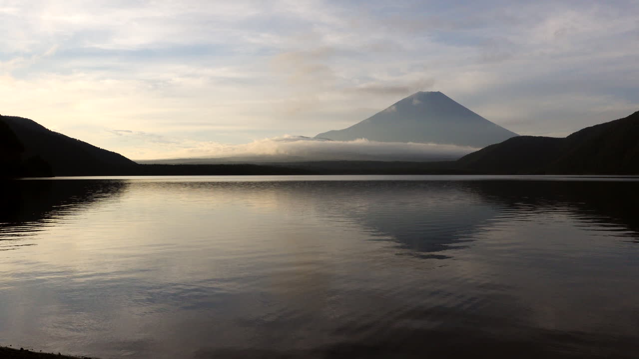 In the soft first light, the camera pans, revealing Lake Motosu's mirror-like surface, mirroring the serene grandeur of Mount Fuji