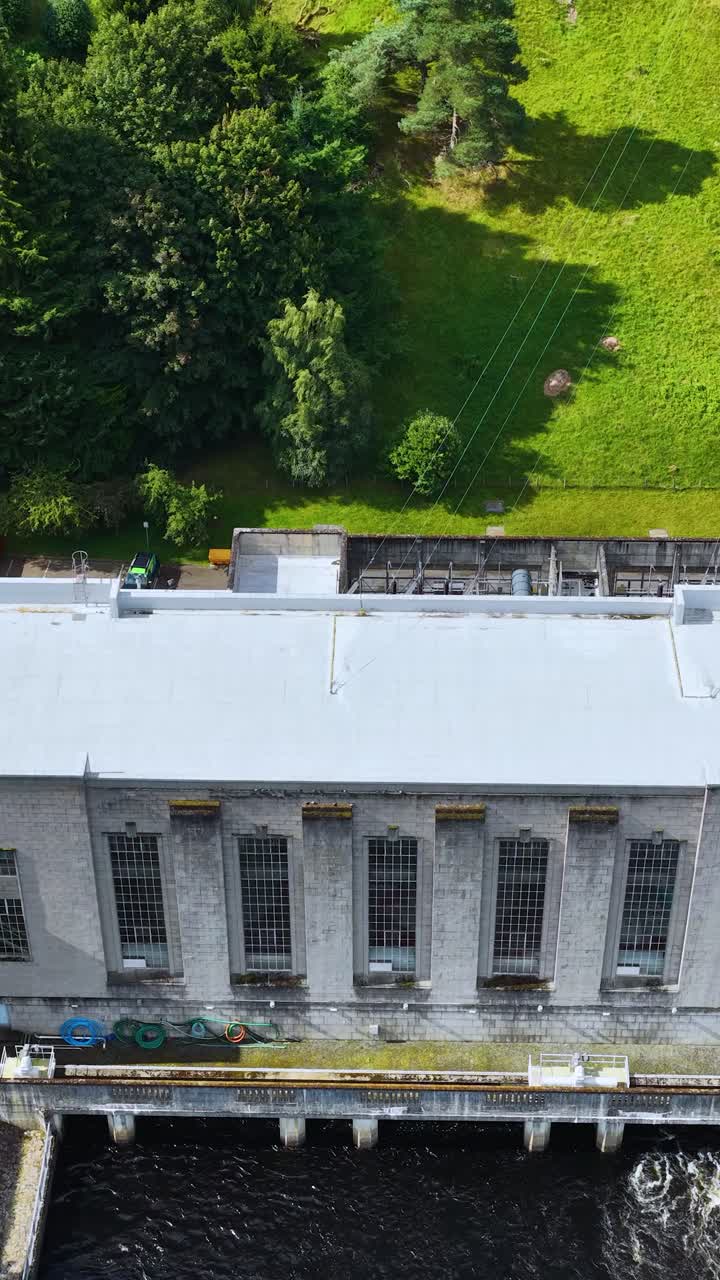 Drone glider over vandkraftværk, flod og grønt landskab i klart dagslys, jævn bevægelse.