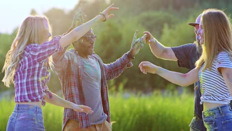 joyful young multiethnic girls and guys celebrating holi festival outdoor in the green field with colorful paints and dances