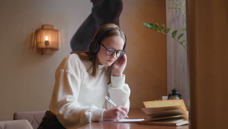 focused lady wearing headset takes notes in notebook with open novel and coffee beside her on wooden desk near window, ambient wall light and potted plant