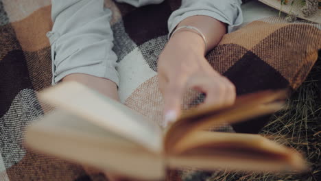 primer plano de una mano de persona con un brazalete, hojeando páginas de libros mientras se acuesta en una manta a cuadros sobre una bala de heno, evocando un entorno de lectura rústico y pacífico