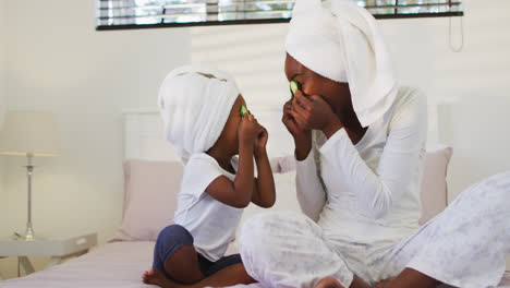 Feliz-Madre-E-Hija-Afroamericana-Usando-Toallas-Sentadas-En-La-Cama-Poniendo-Pepinos-En-Los-Ojos