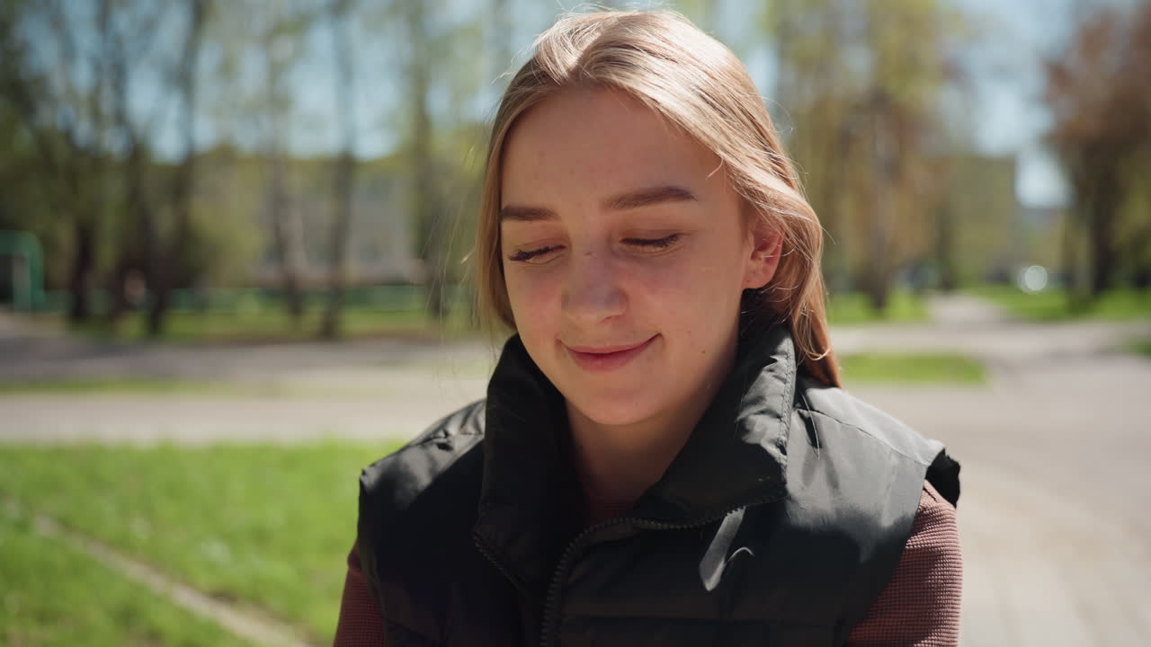 햇빛이 비치는 공원에서 미소 짓고 있는 백인 학생의 클로즈업 사진이에요. 얕은 피사계 심도, 초록색 나무와 통로, 캐주얼한 검은색 조끼와 주근깨가 있으며 라이프스타일 또는 교육 콘텐츠에 어울리는 따뜻하고 친근한 표정을 짓고 있어요.