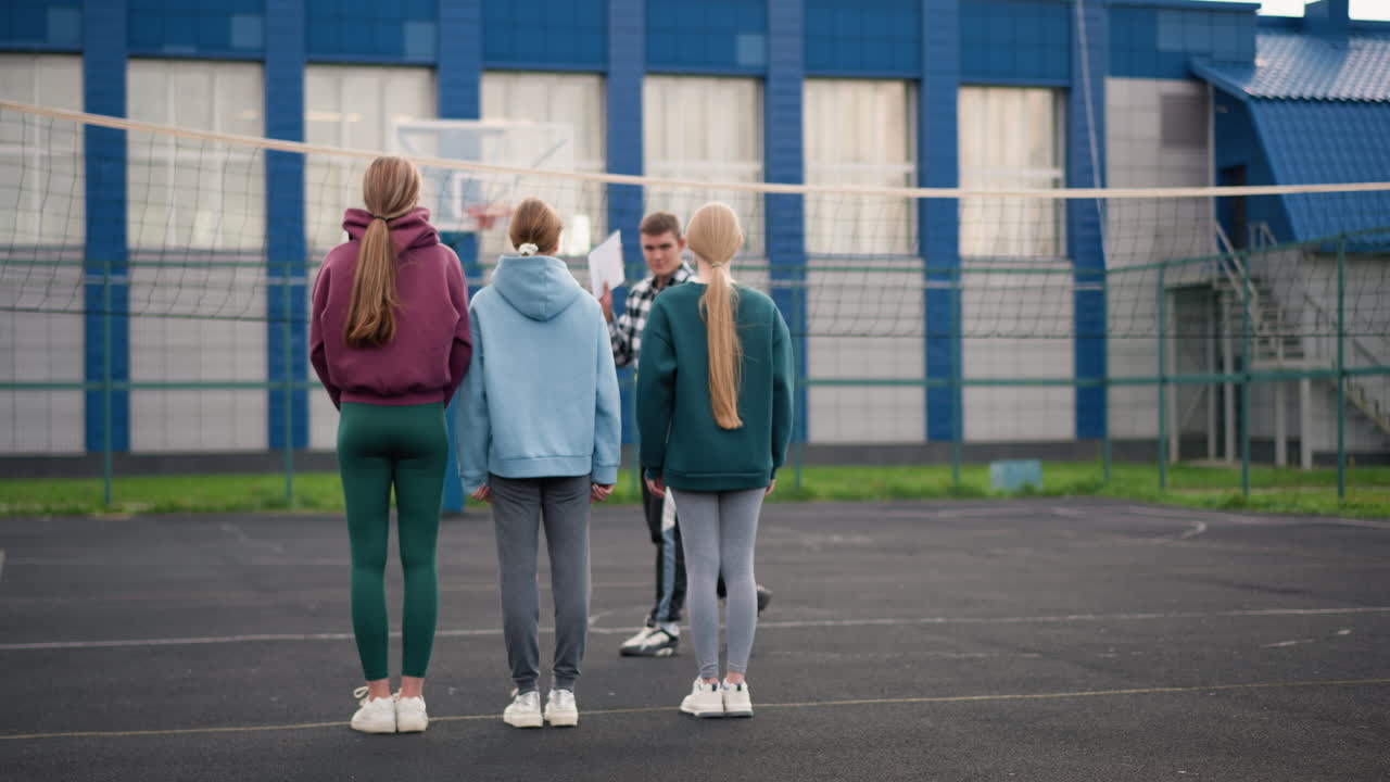 vista trasera de jóvenes atletas con ropa deportiva de pie en la cancha de voleibol, viendo al entrenador rebotar la pelota, su cabello revoloteando en el viento, cancha al aire libre con red, edificio azul y vegetación en el fondo