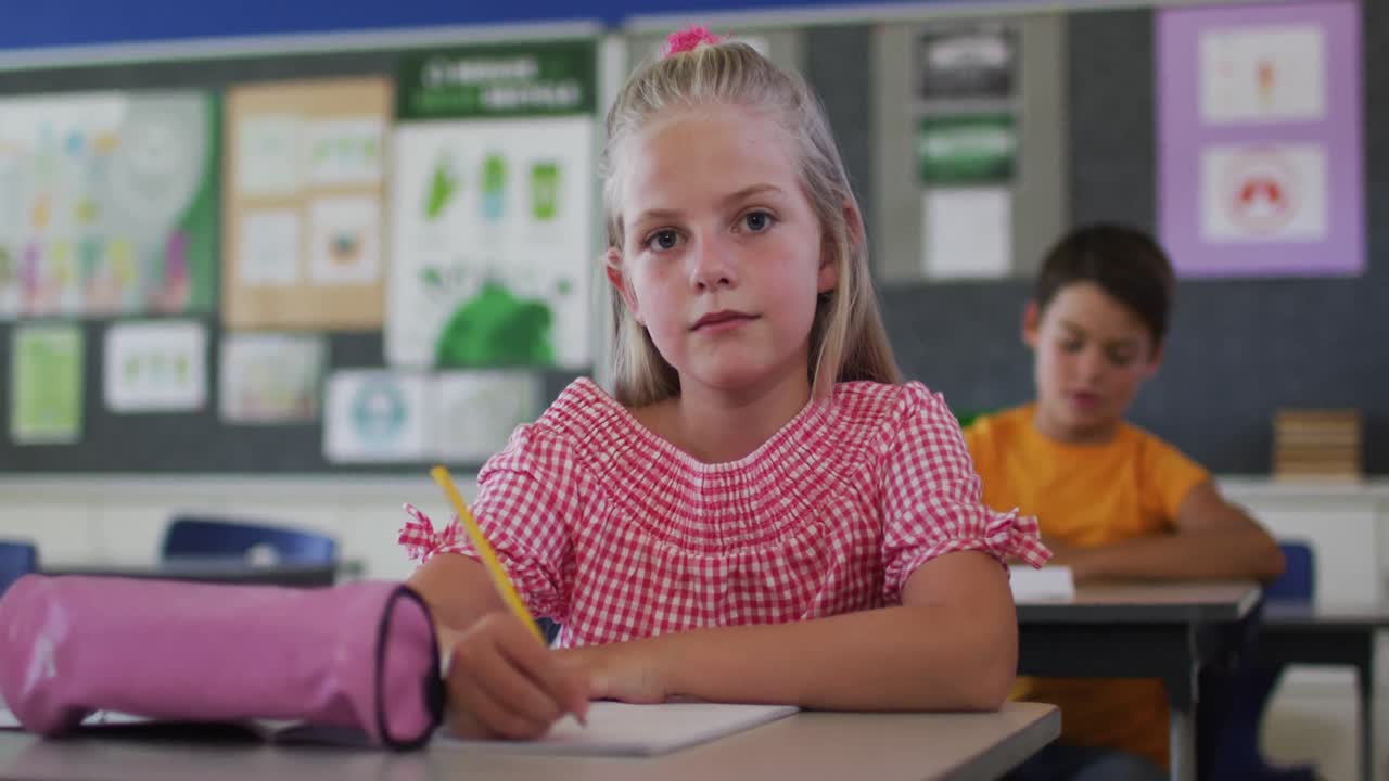 Portrait of happy caucasian schoolgirl sitting at classroom, making notes, looking at camera