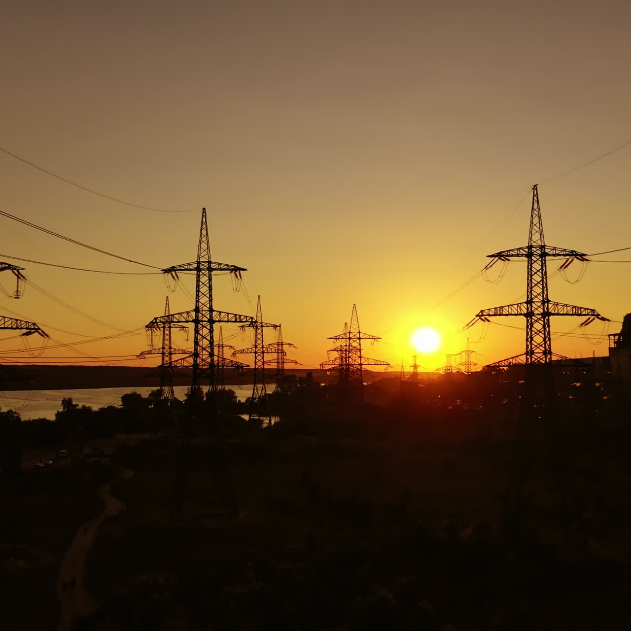 High voltage station at sunset. Tall pylons for electricity transmission. Silhouette of electric towers over sunset time. Camera moving top down.
