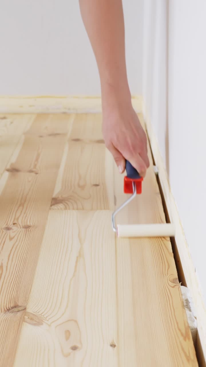 Person applying a transparent finish along the edge of a wooden floor using a small roller near the wall during home renovation, vertical close-up shot