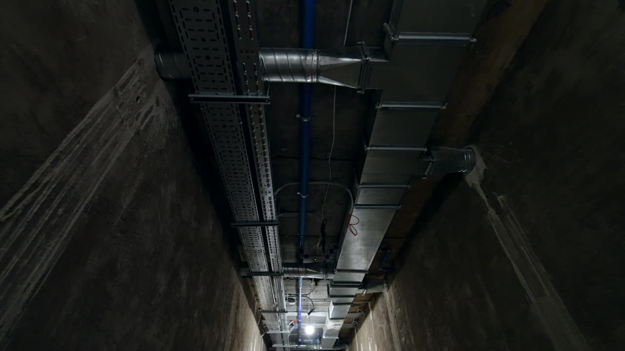 Pipes, wires and metal constructions on the ceiling of the long corridor. Tour by the premises under renovation. Low angle view.