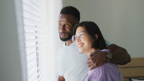 Happy-diverse-couple-embracing-and-looking-through-window-in-bedroom