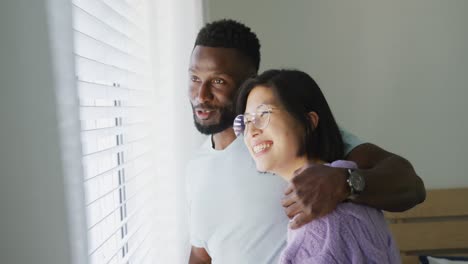 happy diverse couple embracing and looking through window in bedroom