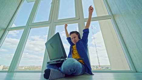 happy boy wins a game on a laptop. joyful teenager in headphones sitting by the window and playing video games on his laptop. gaming concept.