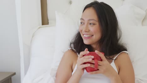 happy asian woman lying in bed and drinking coffee, in slow motion