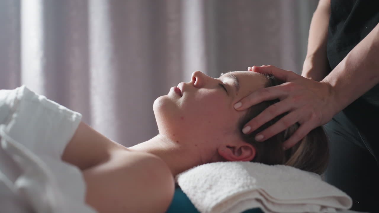 Side view of woman lying on towel-covered bed with eyes closed while therapist gently massages forehead, natural light filtering through gray curtain, creating calm spa setting and sense of relaxation