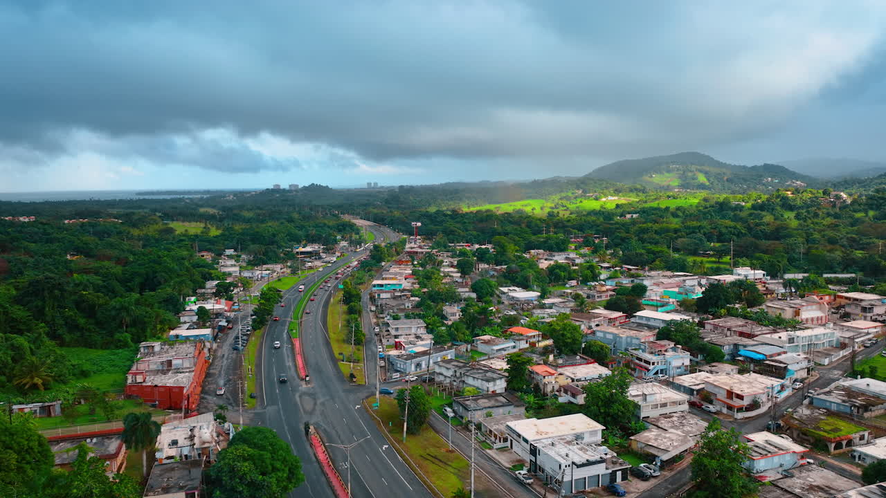 Riprese aeree sopra la strada a quattro corsie lungo il paesaggio urbano a bassa quota. Vegetazione lussureggiante intorno. Un cielo nuvoloso grigio e drammatico copre il cielo. Porto Rico
