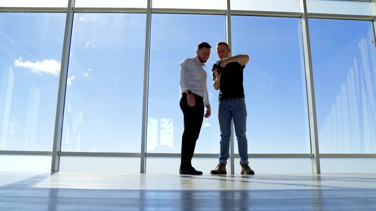 Businessman posing on camera indoors on sky background. Photographer taking photos of a handsome male and showing photos from camera. Panoramic windows view at daytime.