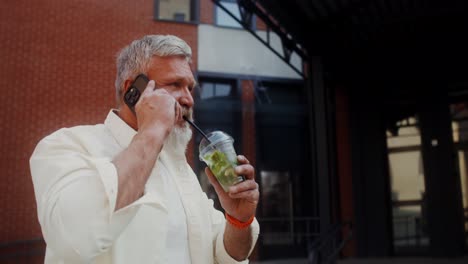 hombre mayor hablando por teléfono y bebiendo una bebida afuera