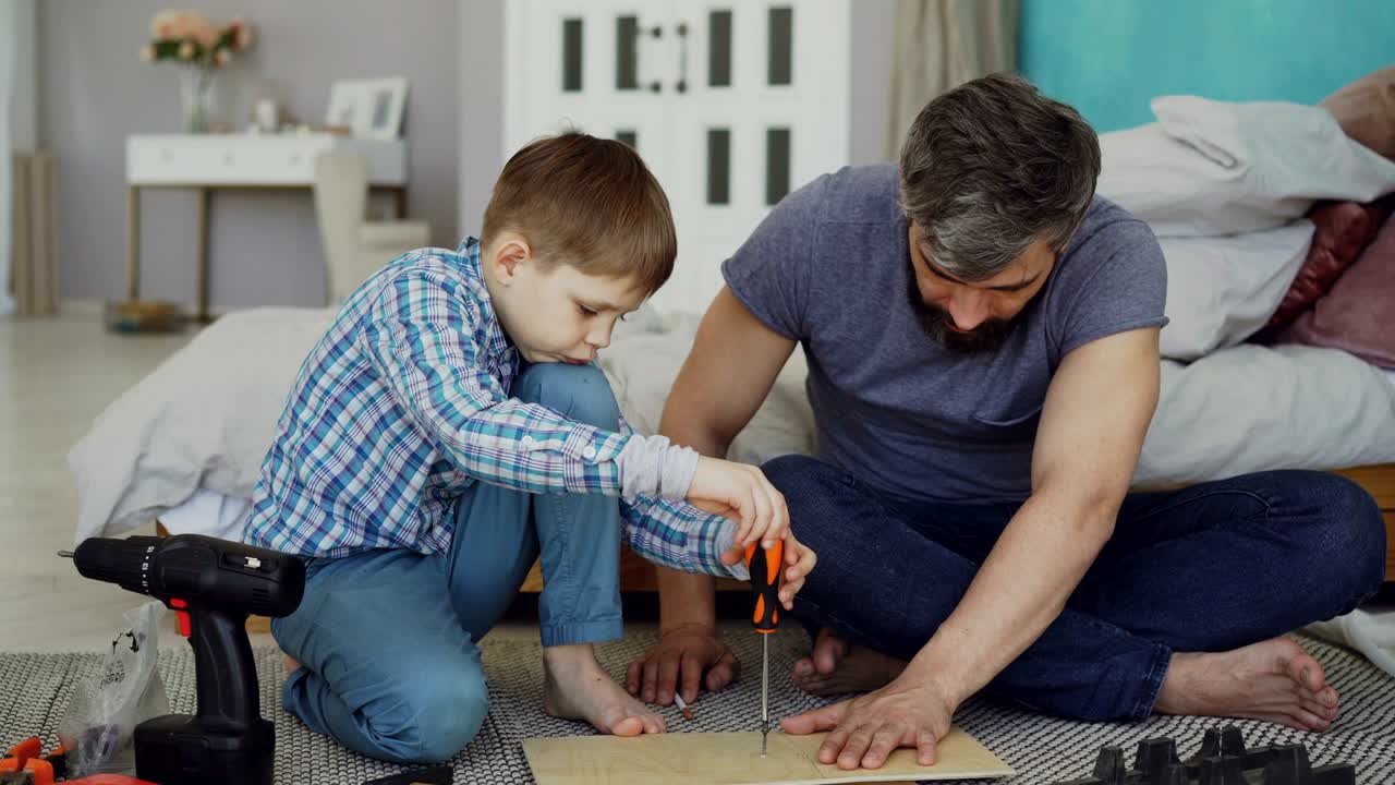 un niño serio se concentra en enroscar pedazos de madera con un destornillador mientras su padre lo ayuda sosteniendo una hoja de madera. infancia y concepto de construcción.