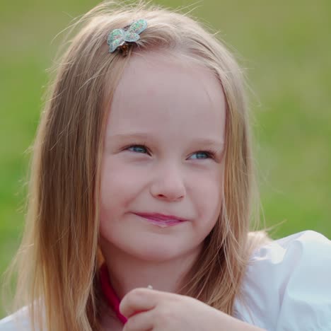 portrait of adorable little girl outdoors. beautiful small blonde child is sitting on the grass and eating on green nature background.