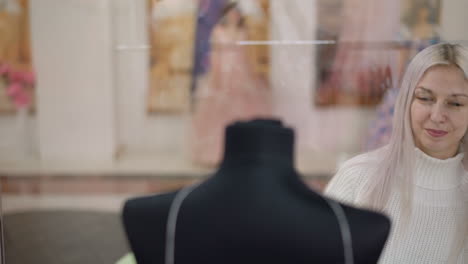 beautiful lady bends down to inspect dinner gown behind glass with admiring smile, walking past mannequins and store decor in mall corridor lit by overhead lights reflecting on glass