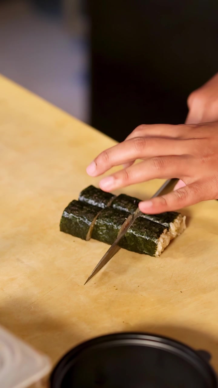 Detailní záběr rukou šéfkuchaře precizně krájících sushi rolky obalené mořskou řasou na dřevěném prkénku